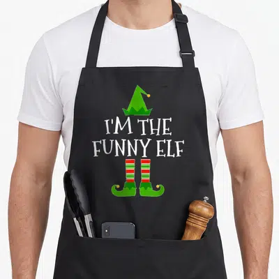 Funny family elf apron - I'M THE FUNNY ELF text with green hat and red striped elf socks — shown on a black cotton canvas apron against a white background