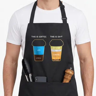 Cappuccino barista coffee apron - Two cartoon cups labeled This Is Coffee and This Is Sh*t showing layered drink — shown on a black cotton canvas apron against a white background