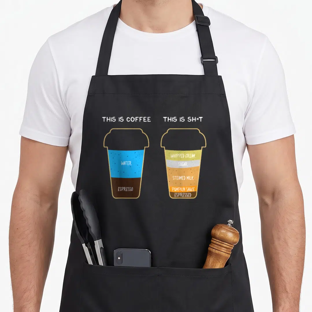 Cappuccino barista coffee apron - Two cartoon cups labeled This Is Coffee and This Is Sh*t showing layered drink — shown on a black cotton canvas apron against a white background