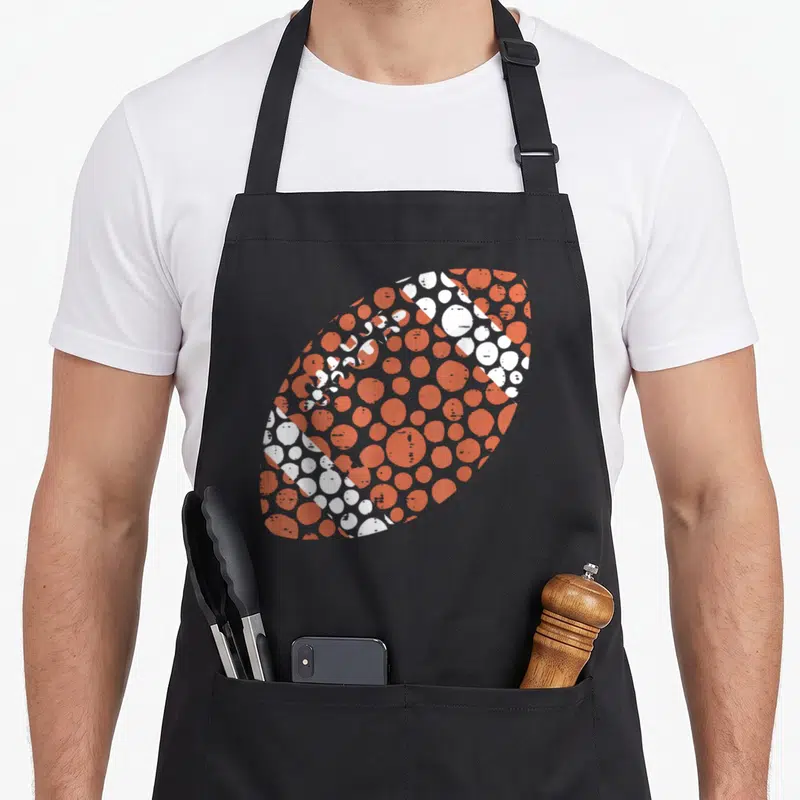 Dot football apron - Oval formed by orange and white dots in minimalist style — shown on a black cotton canvas apron against a white background