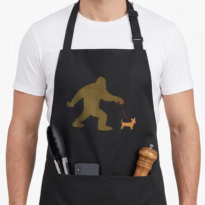 Walking bigfoot apron - Brown Bigfoot silhouette walking a small orange dog on a red leash — shown on a black cotton canvas apron against a white background