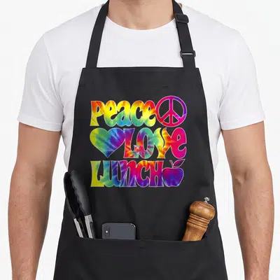Peace lunch lady apron - Tie-dye text reading PEACE LOVE LUNCH with symbols in red yellow blue green purple — shown on a black cotton canvas apron against a white background
