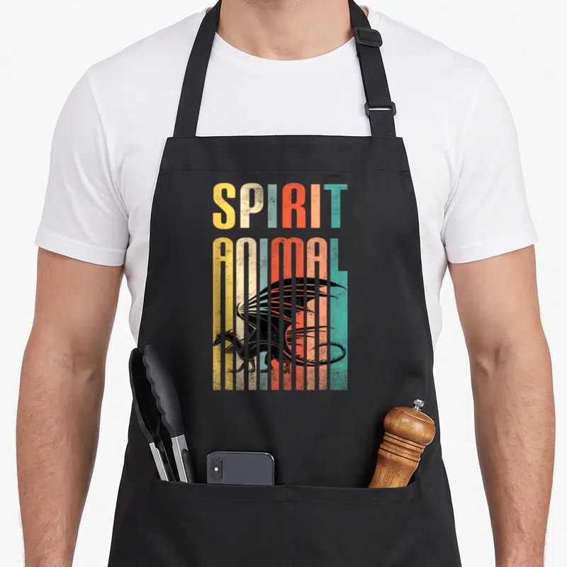 Funny animal dragon apron - Retro SPIRIT ANIMAL text and black dragon behind yellow, orange, red, and teal vertical — shown on a black cotton canvas apron against a white background