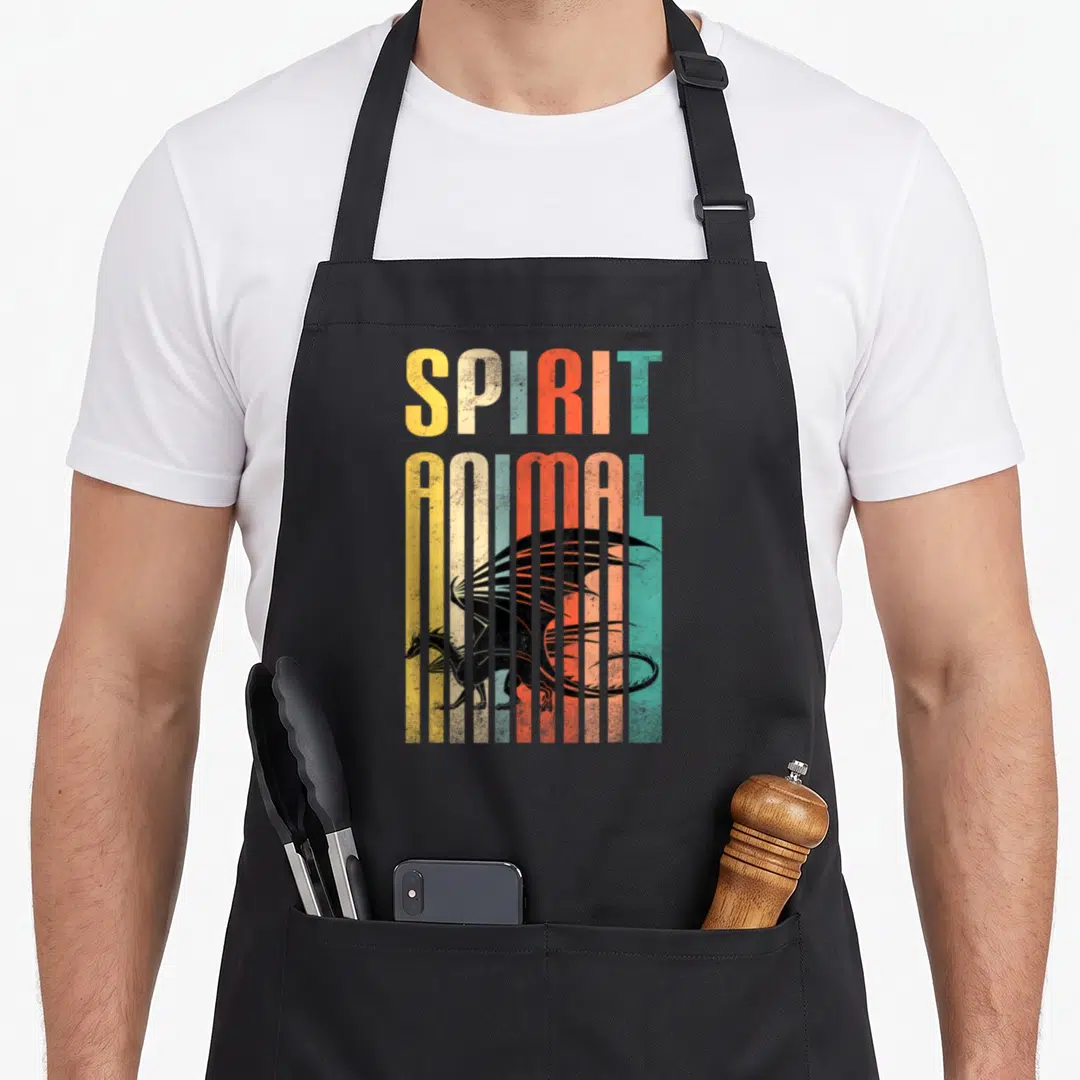 Funny animal dragon apron - Retro SPIRIT ANIMAL text and black dragon behind yellow, orange, red, and teal vertical — shown on a black cotton canvas apron against a white background