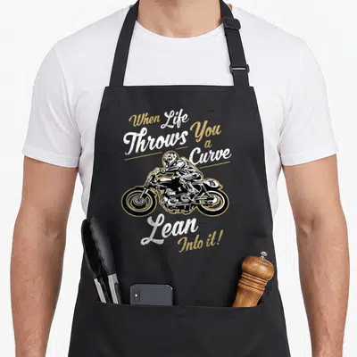 Sarcastic biker apron - Distressed motorcyclist graphic with white and gold text When Life Throws You a Curve Lean — shown on a black cotton canvas apron against a white background