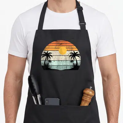 Sunshine summer tree apron - Retro sunset with orange and yellow stripes, two black palm trees, light blue water — shown on a black cotton canvas apron against a white background