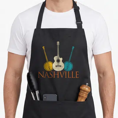 Souvenir country apron - Cream guitar and two banjos in yellow and blue with orange Nashville text below — shown on a black cotton canvas apron against a white background