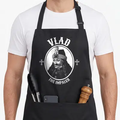 Dracula goth apron - White line art portrait of a man inside a circle with VLAD THE IMPALER text and two crosses on grey — shown on a black cotton canvas apron against a white background