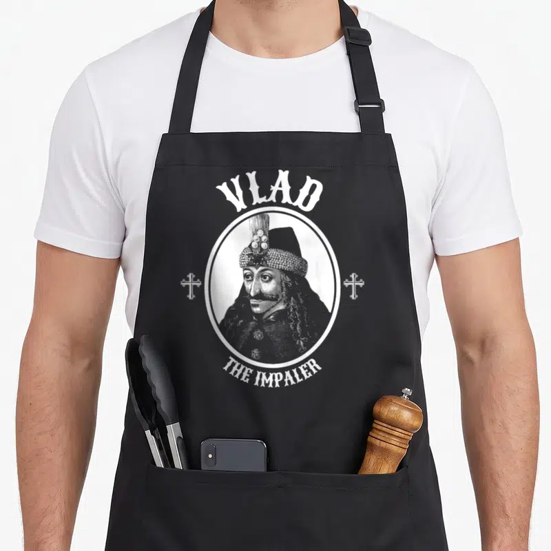 Dracula goth apron - White line art portrait of a man inside a circle with VLAD THE IMPALER text and two crosses on grey — shown on a black cotton canvas apron against a white background