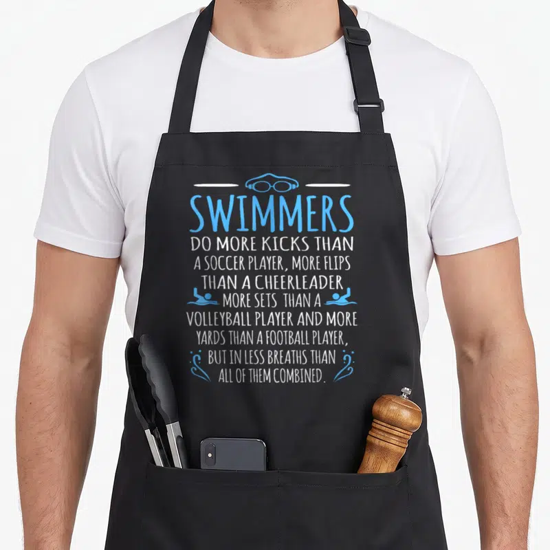 Fact swimming apron - Stacked text reading Swimmers do more kicks than a soccer player with goggles and two swimmer — shown on a black cotton canvas apron against a white background