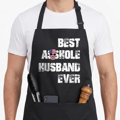 Funny husband apron - Distressed skull with flag pattern overlapping white stacked text reading BEST ASSHOLE HUSBAND — shown on a black cotton canvas apron against a white background