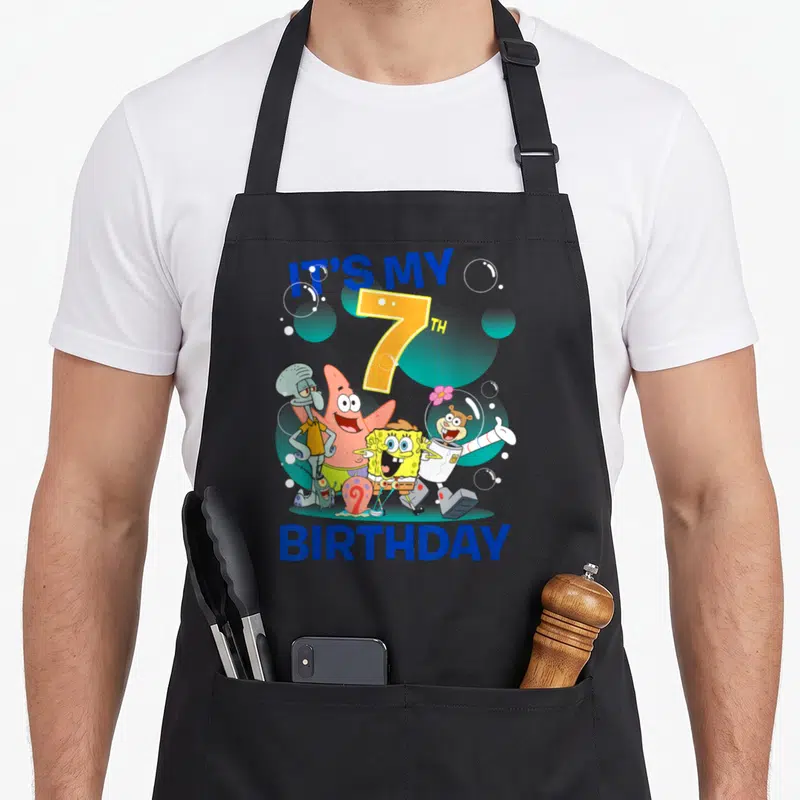 Squarepant 7th birthday apron - Cartoon group of four characters under blue IT'S MY 7TH BIRTHDAY text with bubbles — shown on a black cotton canvas apron against a white background