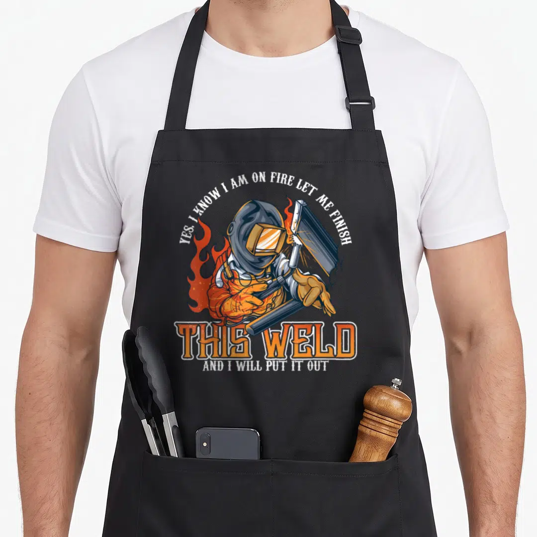 Welder class funny apron - Cartoon welder in orange flames holding torch with arched white text and large orange block — shown on a black cotton canvas apron against a white background