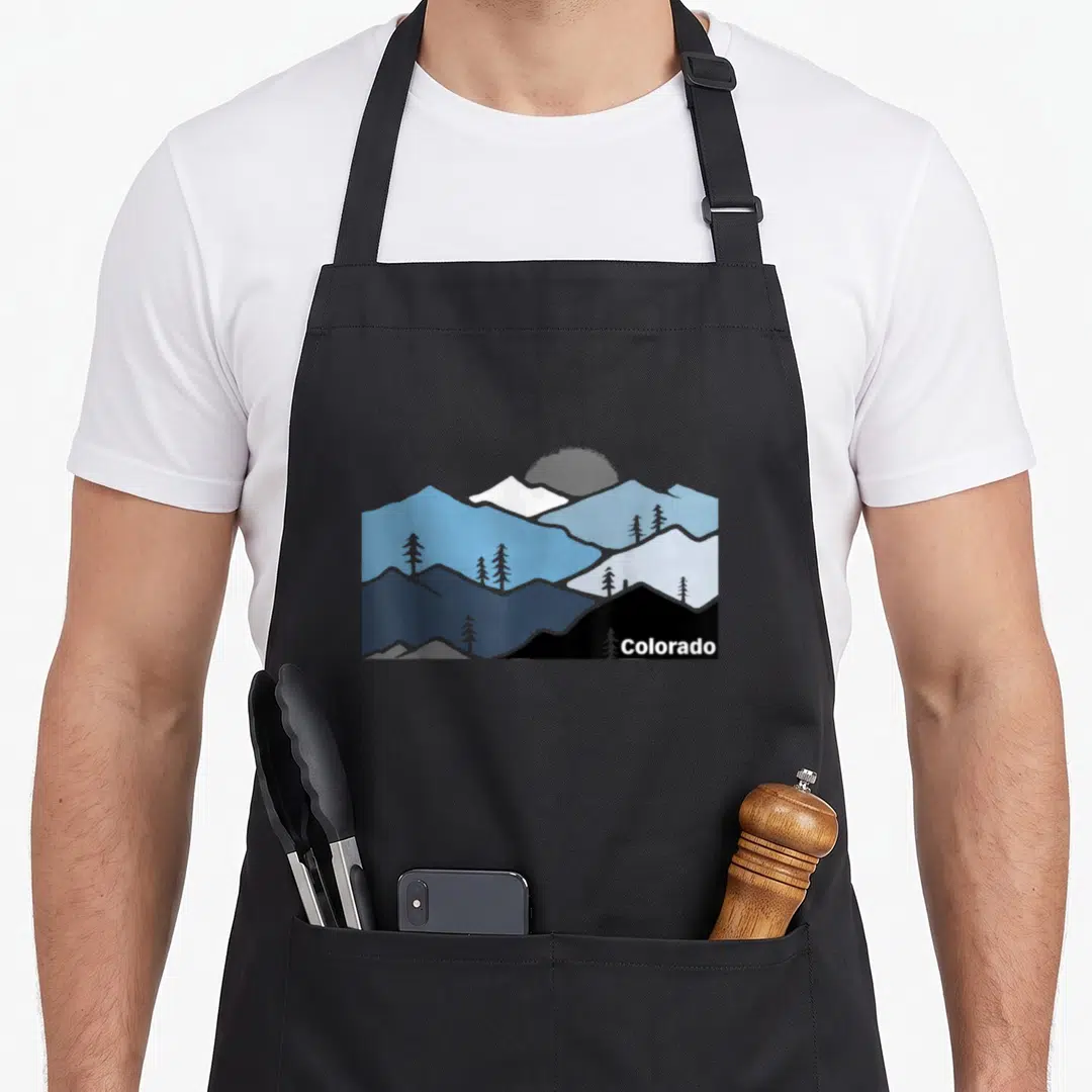 Colorado outdoor apron - Layered blue and grey mountain peaks with pine trees and white Colorado text in bottom right — shown on a black cotton canvas apron against a white background
