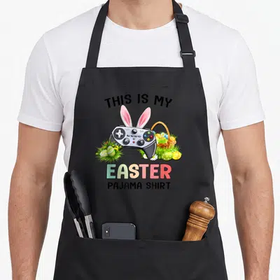 Pajama basket apron - Game controller with pink bunny ears surrounded by Easter eggs, grass, and a basket under printed — shown on a black cotton canvas apron against a white background