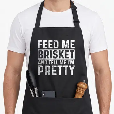 Feed smoker apron - White stacked text reading feed me brisket and tell me i m pretty with a rectangular border around — shown on a black cotton canvas apron against a white background
