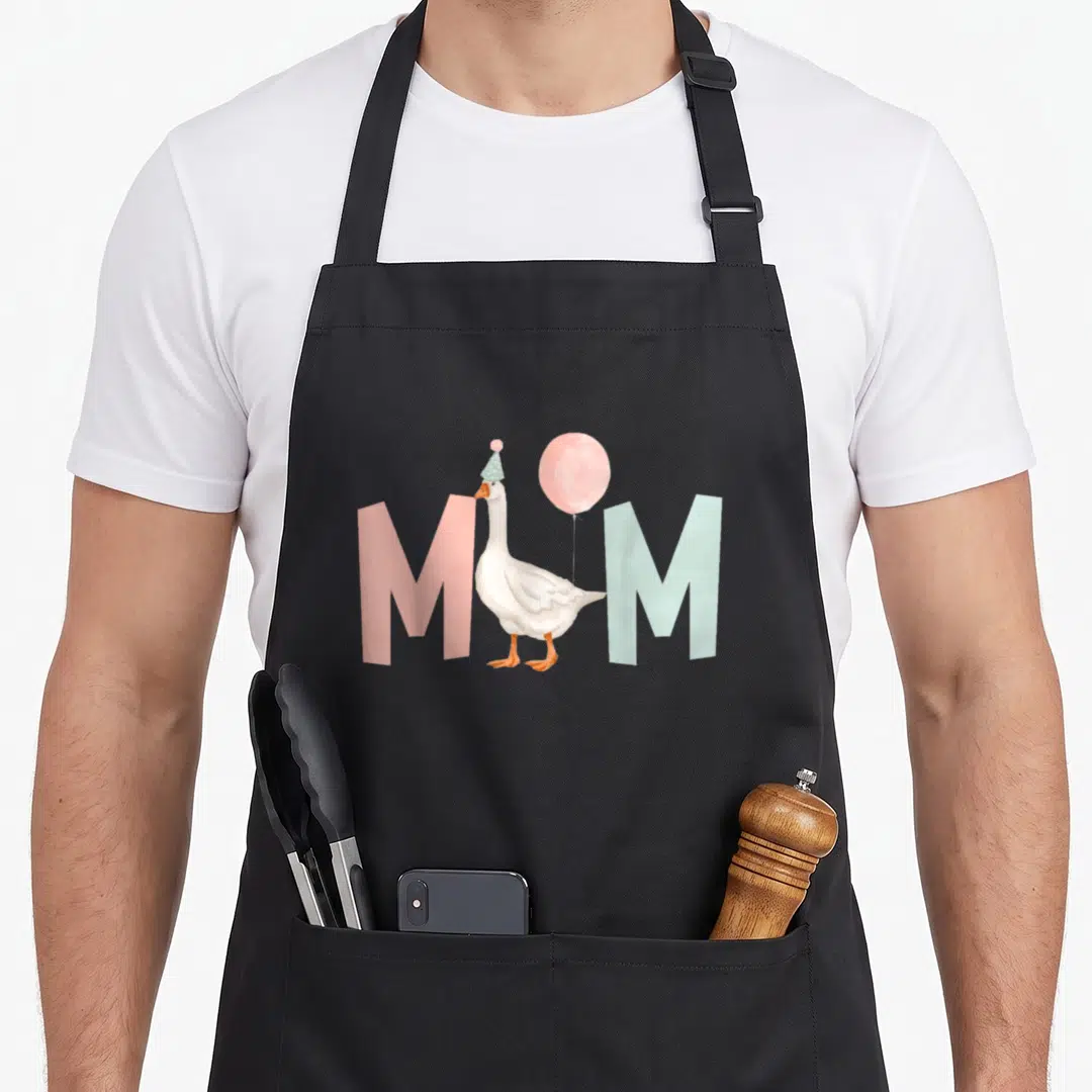Birthday silly apron - White goose with party hat between pink and blue M letters — shown on a black cotton canvas apron against a white background