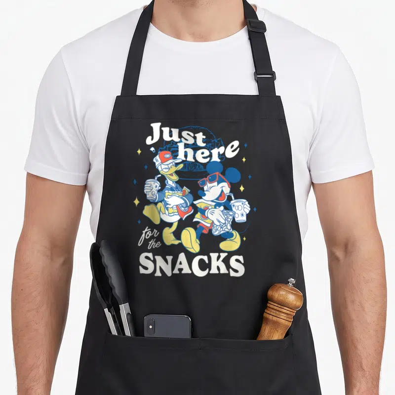 Donald road apron - Mickey and Donald holding snacks with text Just here for the SNACKS in retro colors — shown on a black cotton canvas apron against a white background