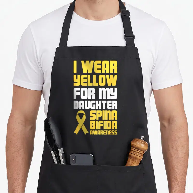 Funny daughter spina bifida awareness apron - Yellow and white text reading I wear yellow for my daughter spina bifida — shown on a black cotton canvas apron against a white background