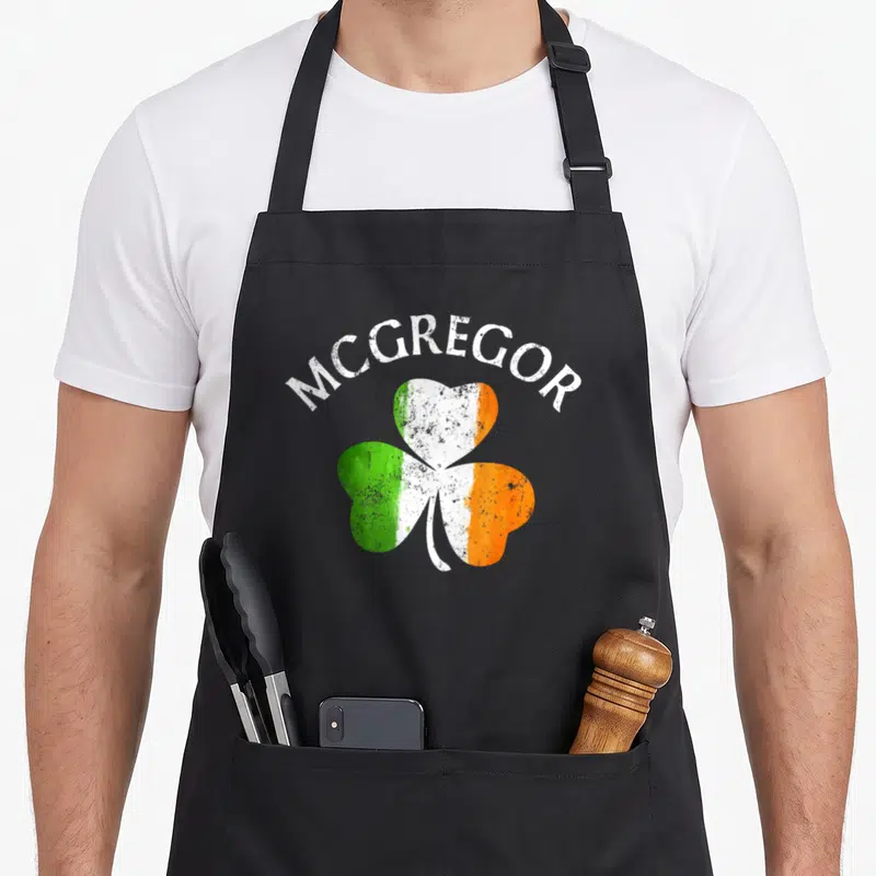 Mcgregor irish aprons - MCGREGOR text arching over a shamrock filled with green white and orange distressed colors — shown on a black cotton canvas apron against a white background