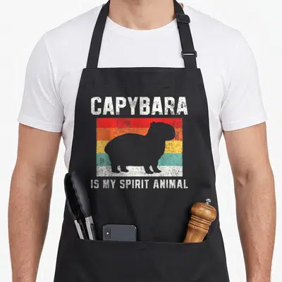 Capybara crew apron - Grey capybara silhouette over five red orange yellow teal white stripes with white text — shown on a black cotton canvas apron against a white background