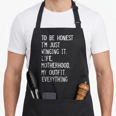 Honest motherhood apron - White stacked text reading TO BE HONEST I'M JUST WINGING IT LIFE MOTHERHOOD MY OUTFIT — shown on a black cotton canvas apron against a white background