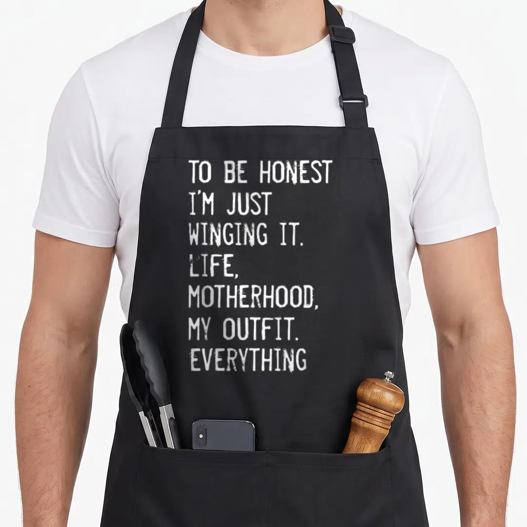 Honest motherhood apron - White stacked text reading TO BE HONEST I'M JUST WINGING IT LIFE MOTHERHOOD MY OUTFIT — shown on a black cotton canvas apron against a white background