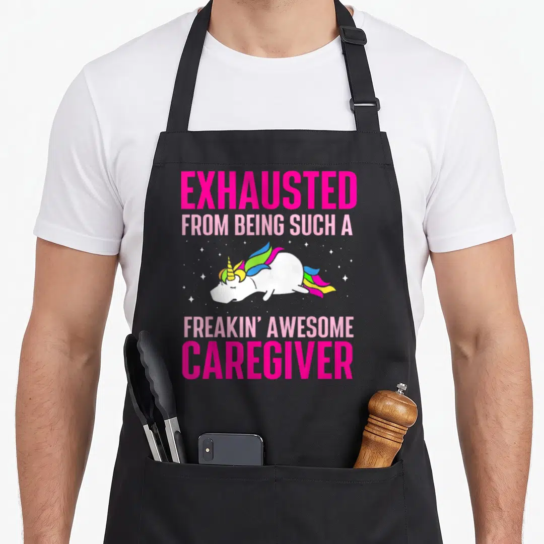 Exhausted caregiver apron - Sleeping white unicorn with rainbow mane below exhausted caregiver text — shown on a black cotton canvas apron against a white background