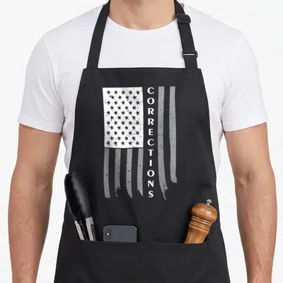 Flag corrections problems apron - Distressed white flag with vertical CORRECTIONS text on black background — shown on a black cotton canvas apron against a white background