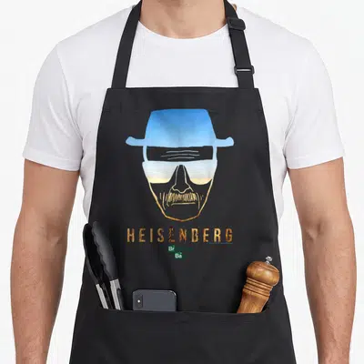 Heisenberg outline of an apron - Stylized face silhouette with hat glasses desert fill gold HEISENBERG text small logo — shown on a black cotton canvas apron against a white background