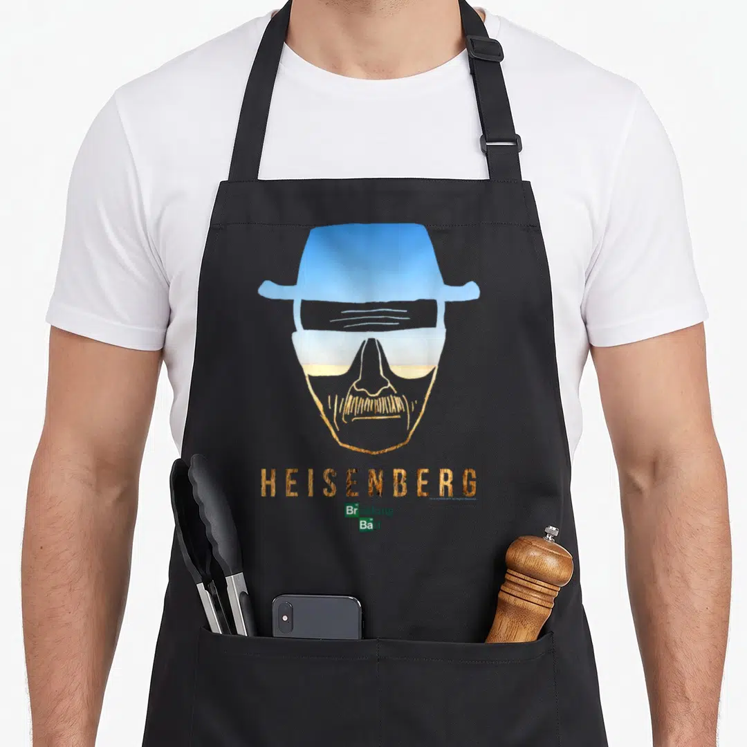 Heisenberg outline of an apron - Stylized face silhouette with hat glasses desert fill gold HEISENBERG text small logo — shown on a black cotton canvas apron against a white background