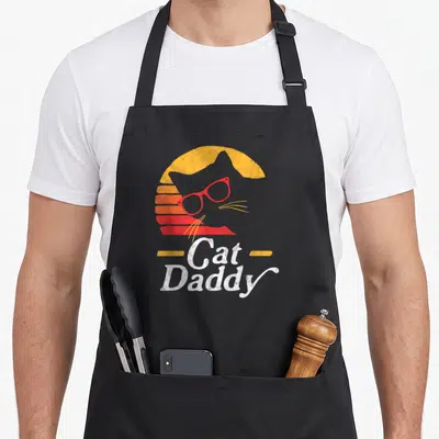 Hipster cat apron - Grey cat head wearing red glasses in front of a yellow and red striped sun with Cat Daddy text — shown on a black cotton canvas apron against a white background
