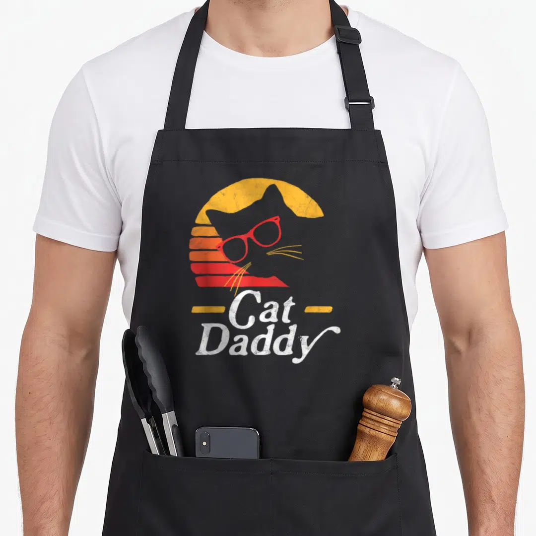 Hipster cat apron - Grey cat head wearing red glasses in front of a yellow and red striped sun with Cat Daddy text — shown on a black cotton canvas apron against a white background