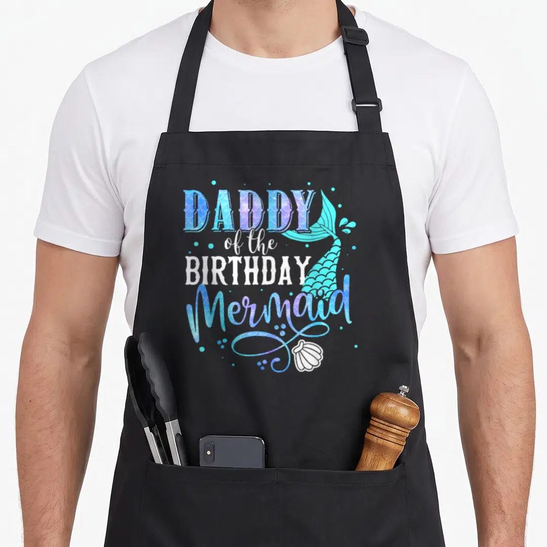 Mermaid grill daddy apron - Stacked DADDY of the BIRTHDAY Mermaid text with teal tail and seashell in blue purple teal — shown on a black cotton canvas apron against a white background