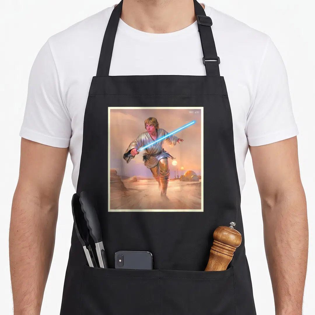 Vintage worlds greatest apron - Luke Skywalker running with blue lightsaber on beige desert with TRUE JEDI text — shown on a black cotton canvas apron against a white background