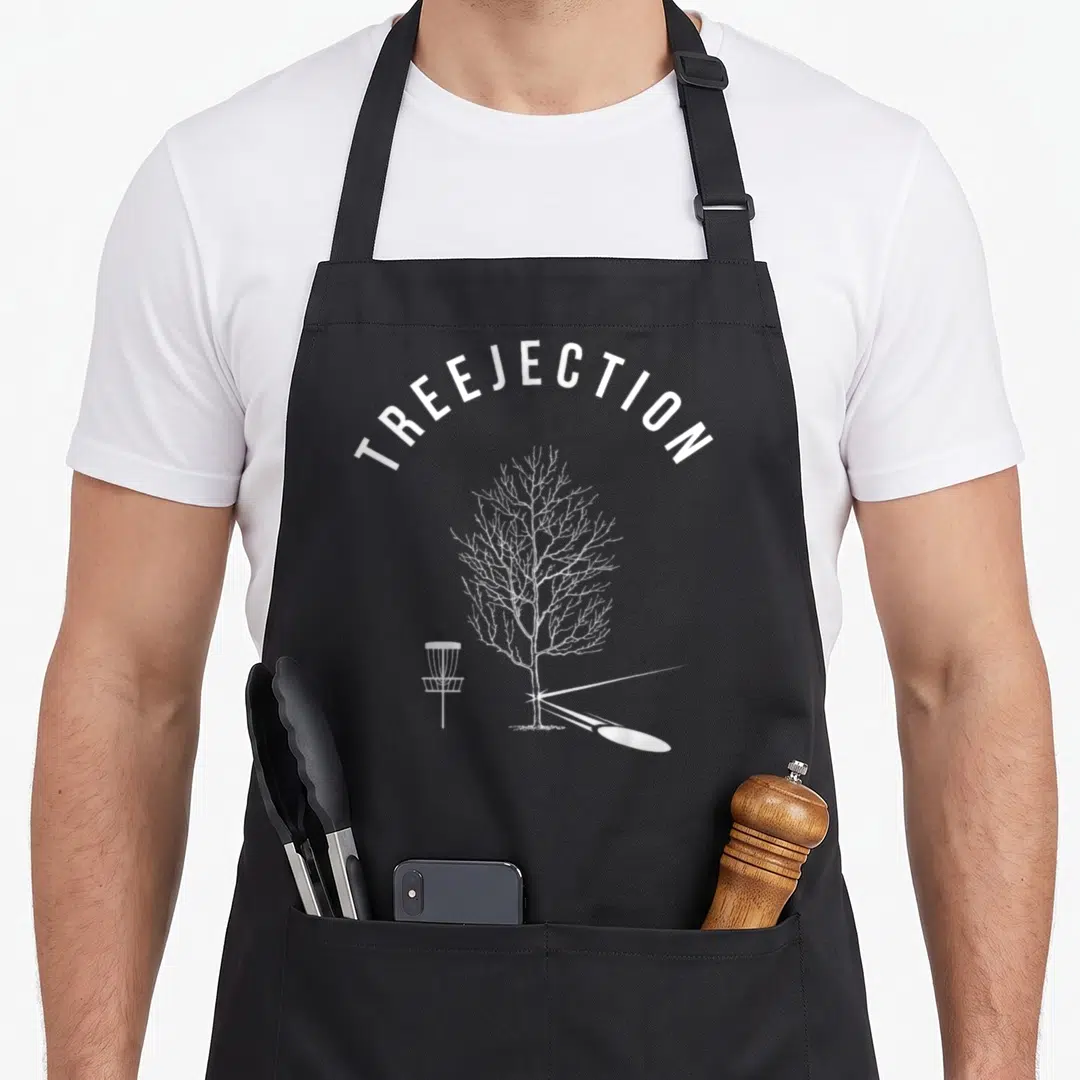 Treejection golf apron - White arched TREEJECTION text above a leafless tree, disc golf basket, and bouncing disc — shown on a black cotton canvas apron against a white background