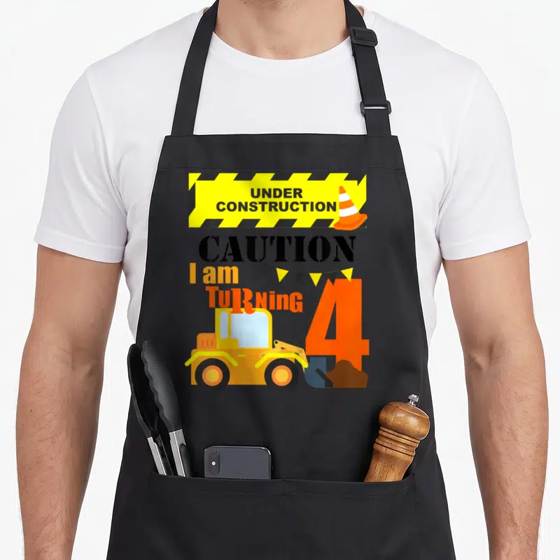 Age truck apron - Yellow construction banner with cone, I am turning 4 text, and loader scooping dirt — shown on a black cotton canvas apron against a white background