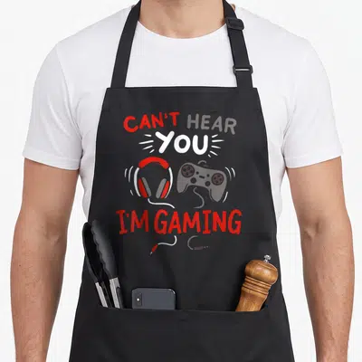 Funny hear gaming apron - Can't Hear You I'm Gaming text with red white grey headphones and controller cartoon art — shown on a black cotton canvas apron against a white background