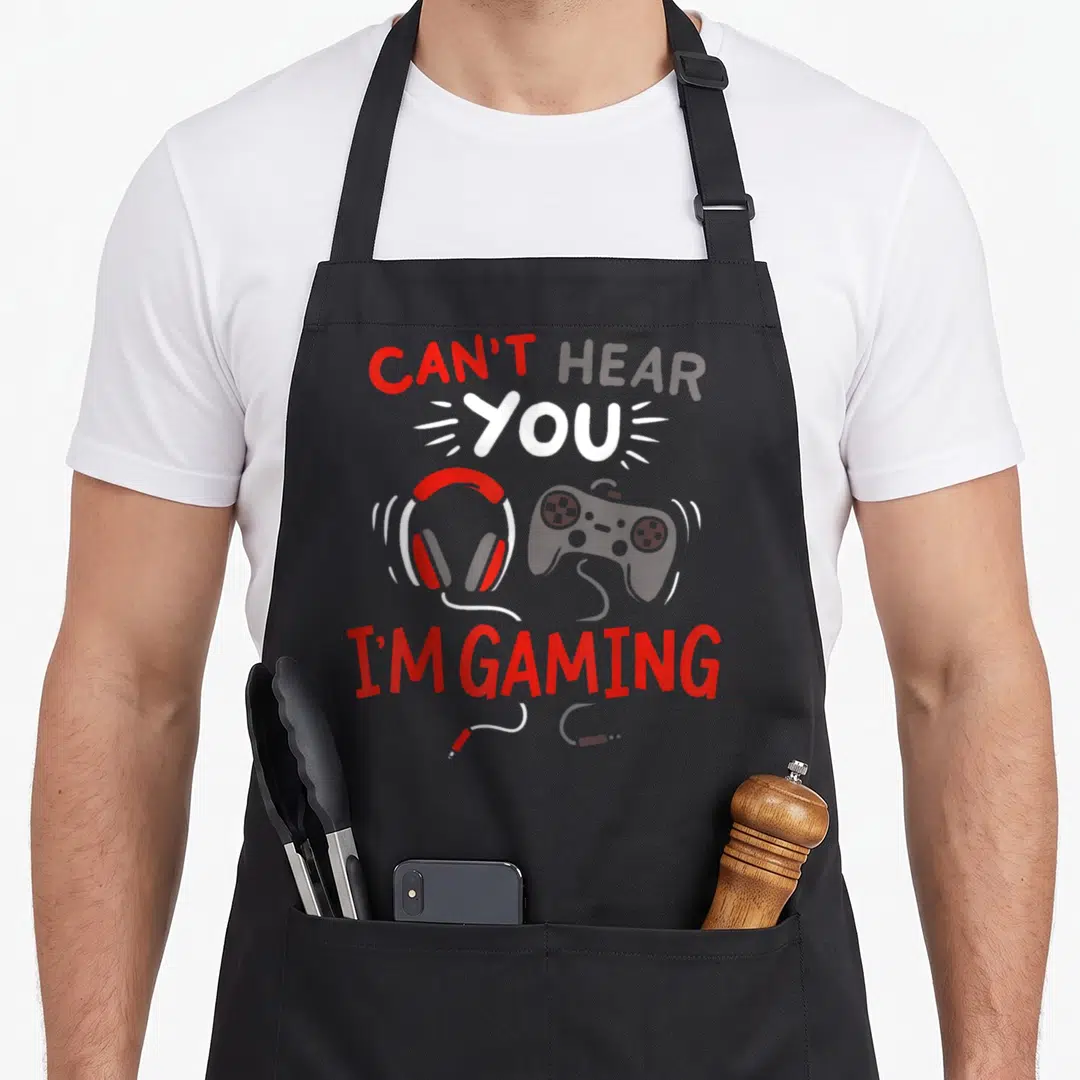 Funny hear gaming apron - Can't Hear You I'm Gaming text with red white grey headphones and controller cartoon art — shown on a black cotton canvas apron against a white background