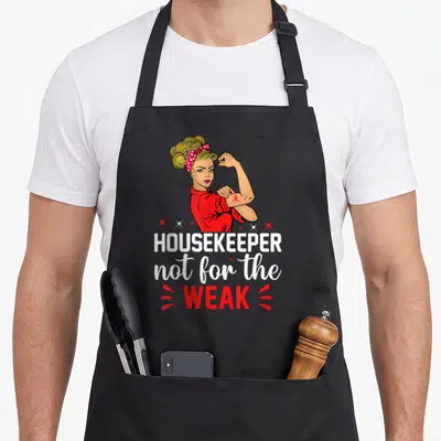 Cleaning housekeeping housekeeper apron - Cartoon woman flexing arm above HOUSEKEEPER not for the WEAK text — shown on a black cotton canvas apron against a white background