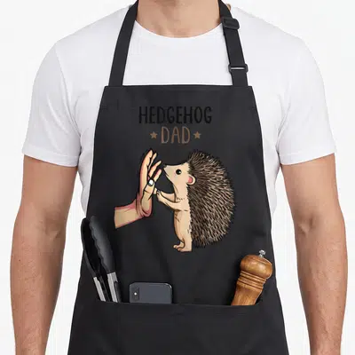 Cute hedgehog dad apron - Cartoon hedgehog high-fiving human hand under stacked HEDGEHOG DAD text in brown tan peach — shown on a black cotton canvas apron against a white background