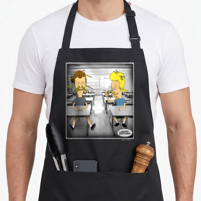 Beavis pastitsio apron - Two cartoon boys at desks with pencils and scissors in hair, grey classroom background — shown on a black cotton canvas apron against a white background