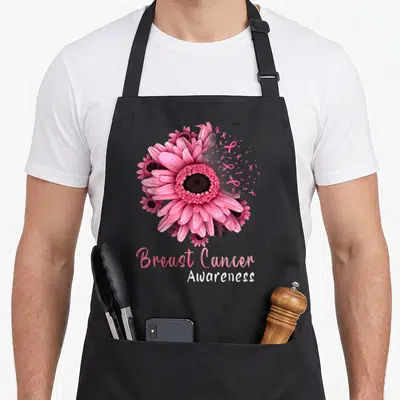 Sunflower ribbon apron - Pink gerbera daisy with floating ribbons and Breast Cancer Awareness text — shown on a black cotton canvas apron against a white background