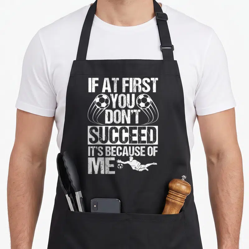 Goalie sports team aprons - White stacked text reading IF AT FIRST YOU DON'T SUCCEED IT'S BECAUSE OF ME with two soccer — shown on a black cotton canvas apron against a white background