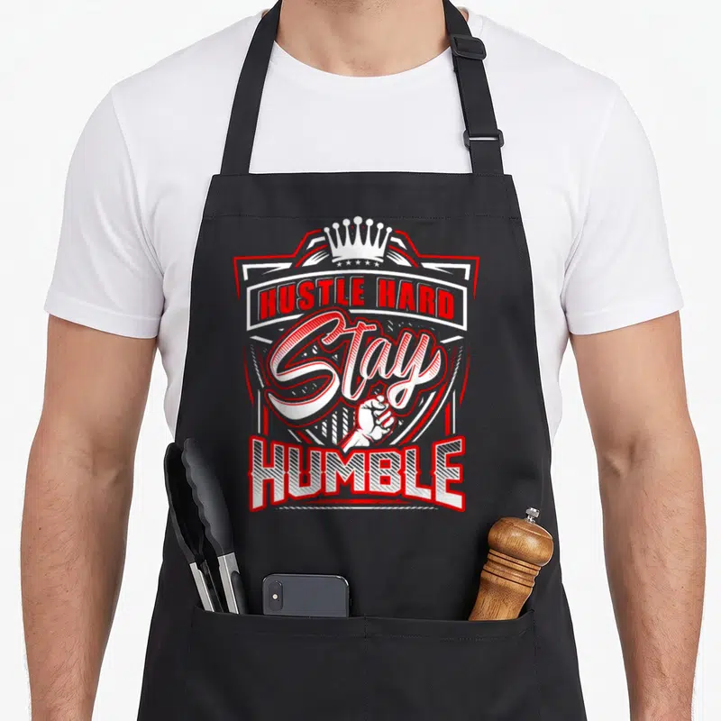 Hustle urban apron - Red and white shield with crown, fist, and HUSTLE HARD Stay HUMBLE text — shown on a black cotton canvas apron against a white background