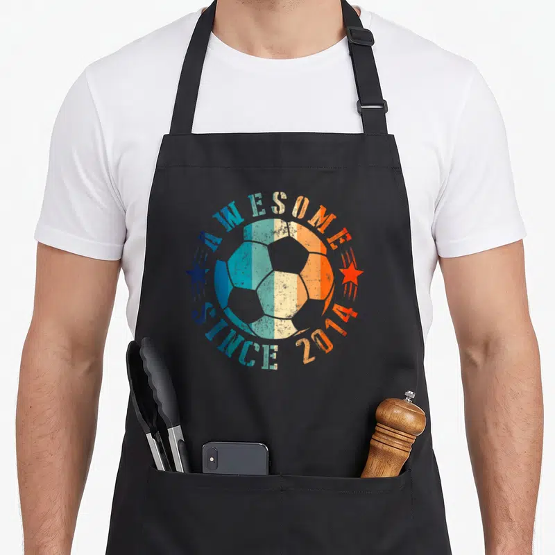 9th soccer birthday apron - Retro soccer ball with Awesome Since 2014 text and two stars in blue teal beige and orange — shown on a black cotton canvas apron against a white background