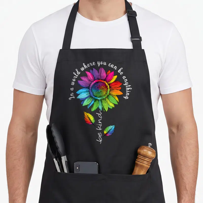 Lgbtq sunflower apron - Rainbow sunflower with arched text and two rainbow leaves flanking vertical be kind script — shown on a black cotton canvas apron against a white background
