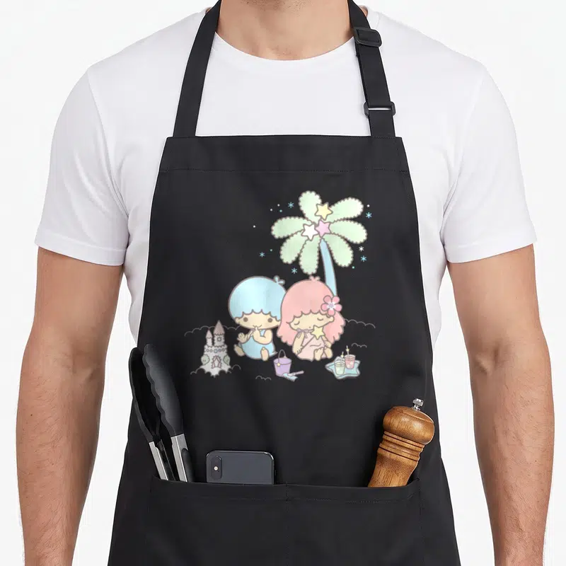 Twin summer aprons - Two cartoon children in pastel clothes sitting by a sandcastle under a palm tree decorated — shown on a black cotton canvas apron against a white background