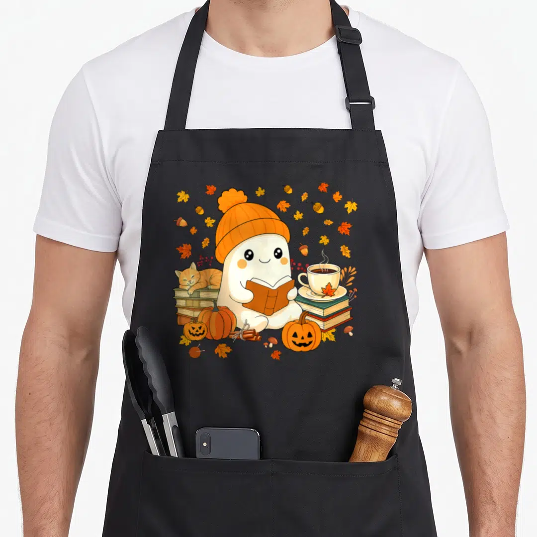 Ghost fall aprons - Cartoon ghost in orange beanie reading book surrounded by autumn leaves, pumpkins, and sleeping cat — shown on a black cotton canvas apron against a white background