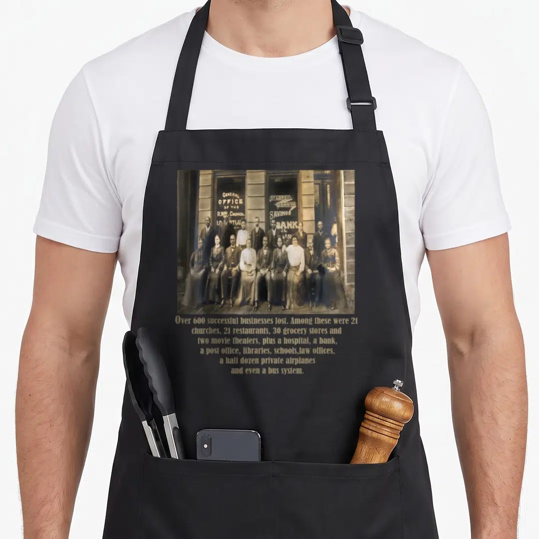 Greenwood tulsa apron - Sepia group photo above gold text listing 600 lost businesses like banks and theaters — shown on a black cotton canvas apron against a white background